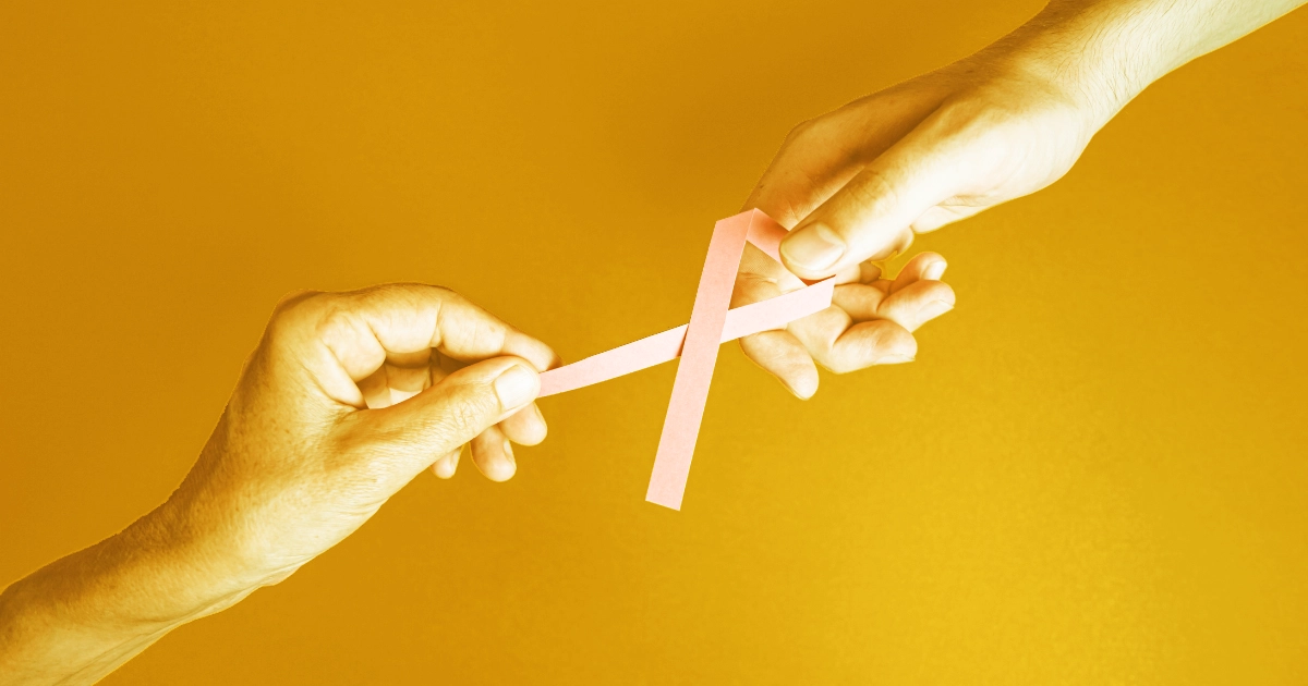 One woman's hand and one man's hand with a Pink Cancer treatment bow being handed to and also held by the other hand